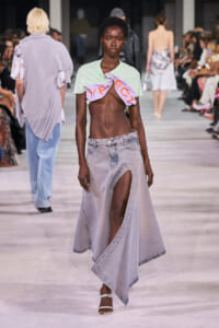 Model on a runway wearing a mint cropped top twisted with a lavender cloth, paired with a high-slit light denim skirt and white sandals; audience watches.