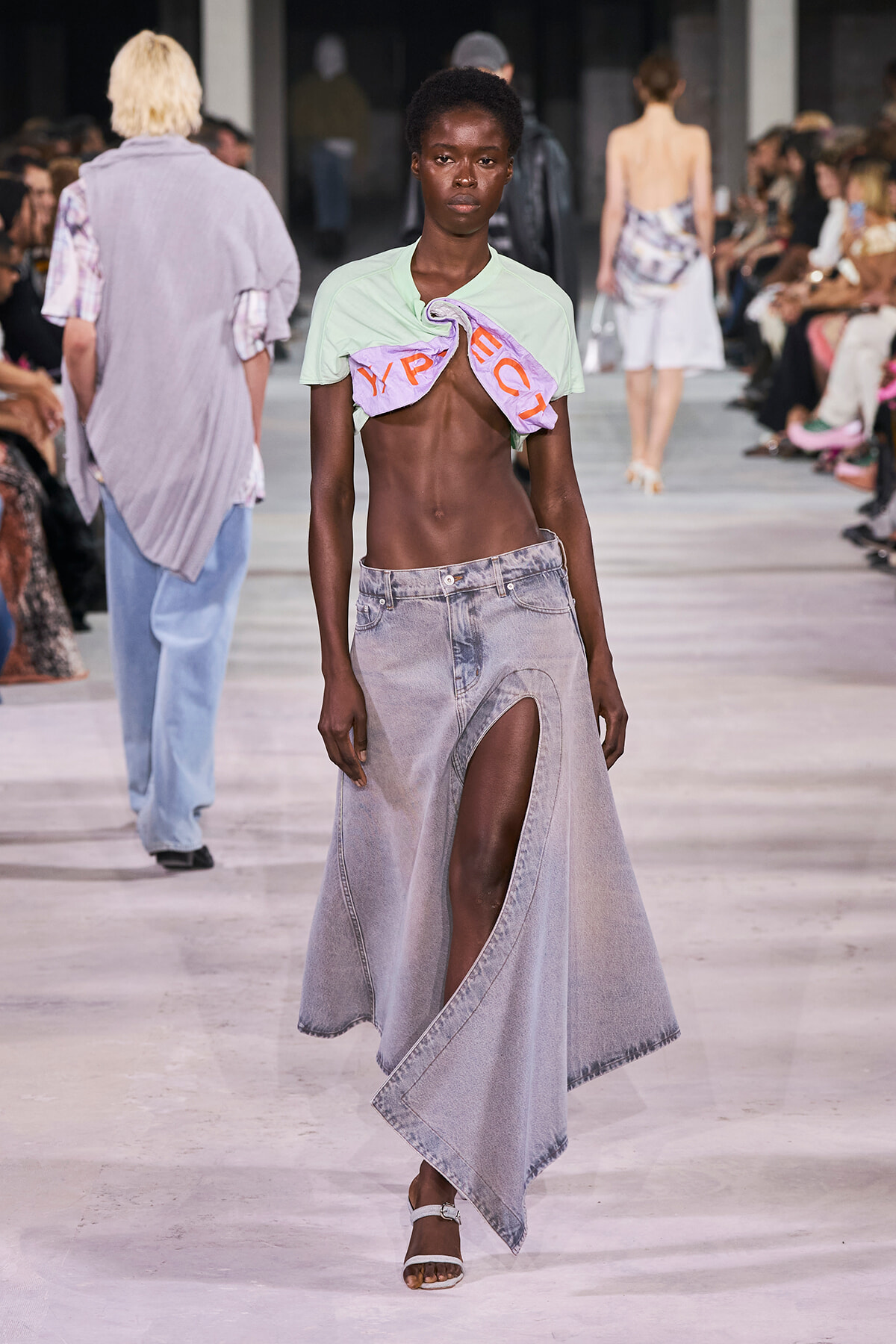 Model on a runway wearing a mint cropped top twisted with a lavender cloth, paired with a high-slit light denim skirt and white sandals; audience watches.