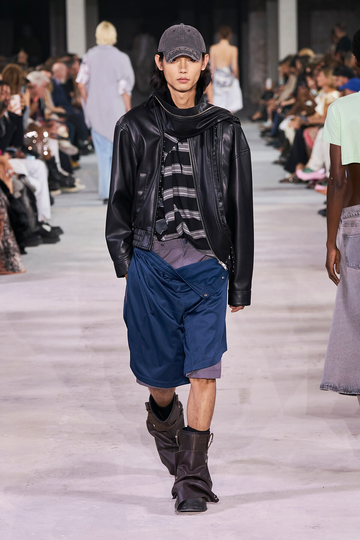 Male model walks the runway in a black leather jacket, striped shirt, blue shorts, gray cap, and brown boots with spectators in the background.
