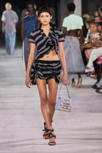Model on a runway wearing a black and gray striped knotted crop top with a matching wrapped mini skirt, carrying a light blue printed handbag.