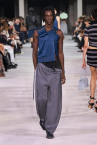 Model walks the runway in a blue sleeveless top with a gathered detail paired with oversized gray sweatpants and black boots, audience in background.