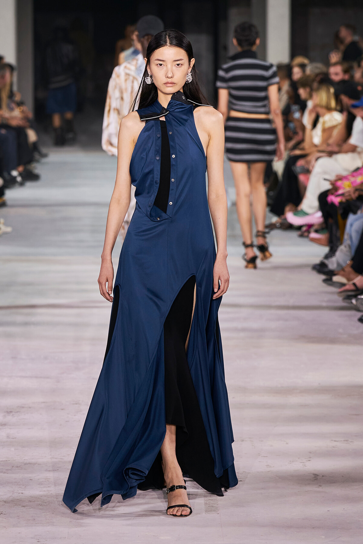 Model walks a runway in a navy blue halter dress with a high slit, audience visible on both sides of the catwalk.