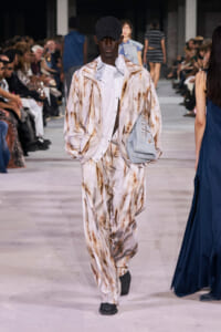 Runway model in a beige-brown marble-pattern suit with a white shirt, black cap, and a light denim bag slung over the shoulder.