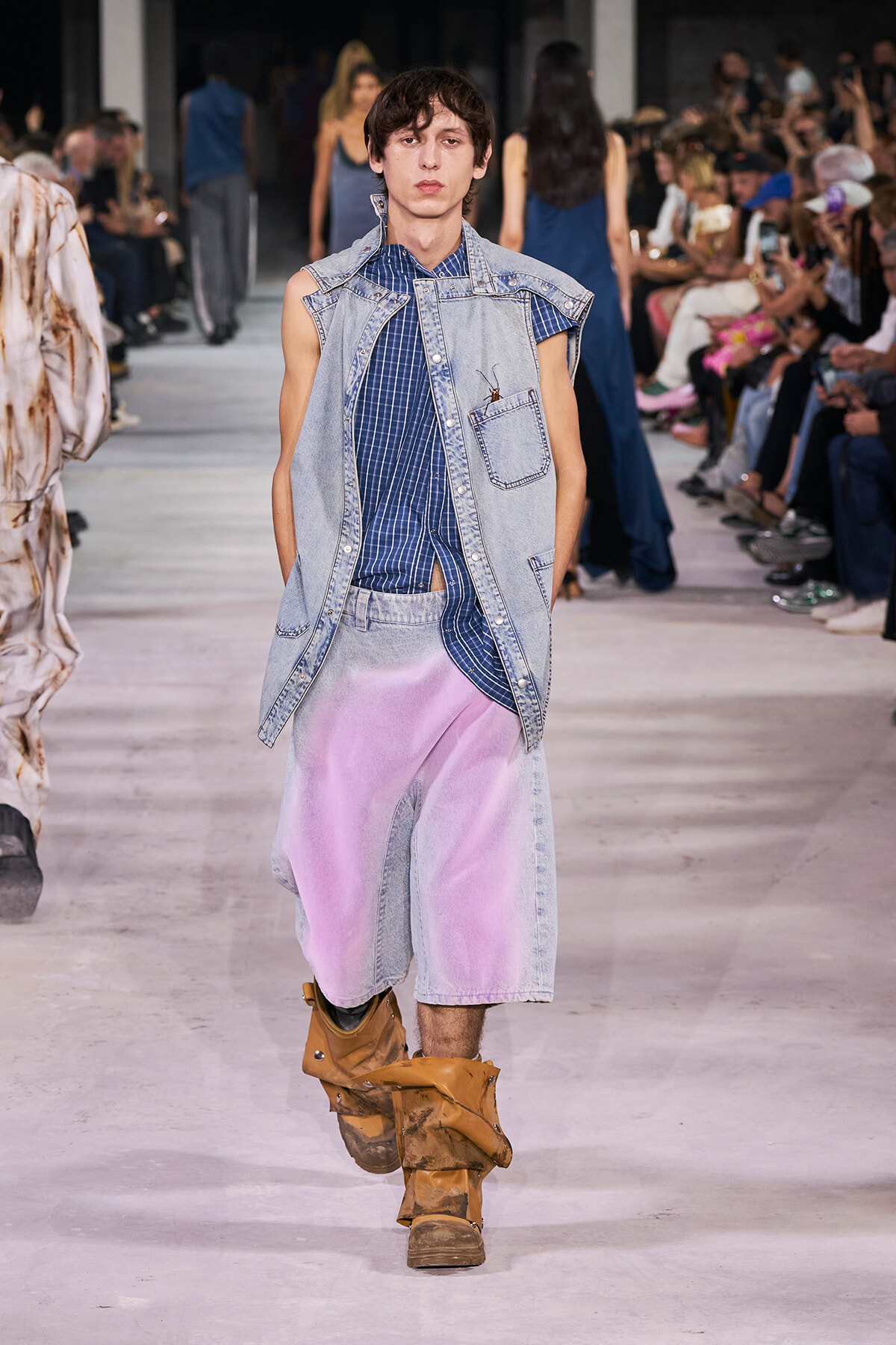 Male model walks a fashion runway in a light denim sleeveless vest over a blue plaid shirt, lavender denim shorts, and tan boots; seated audience in background.