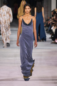 Female model walks the runway in a long lavender velvet dress with a deep neckline, audience visible on sides.
