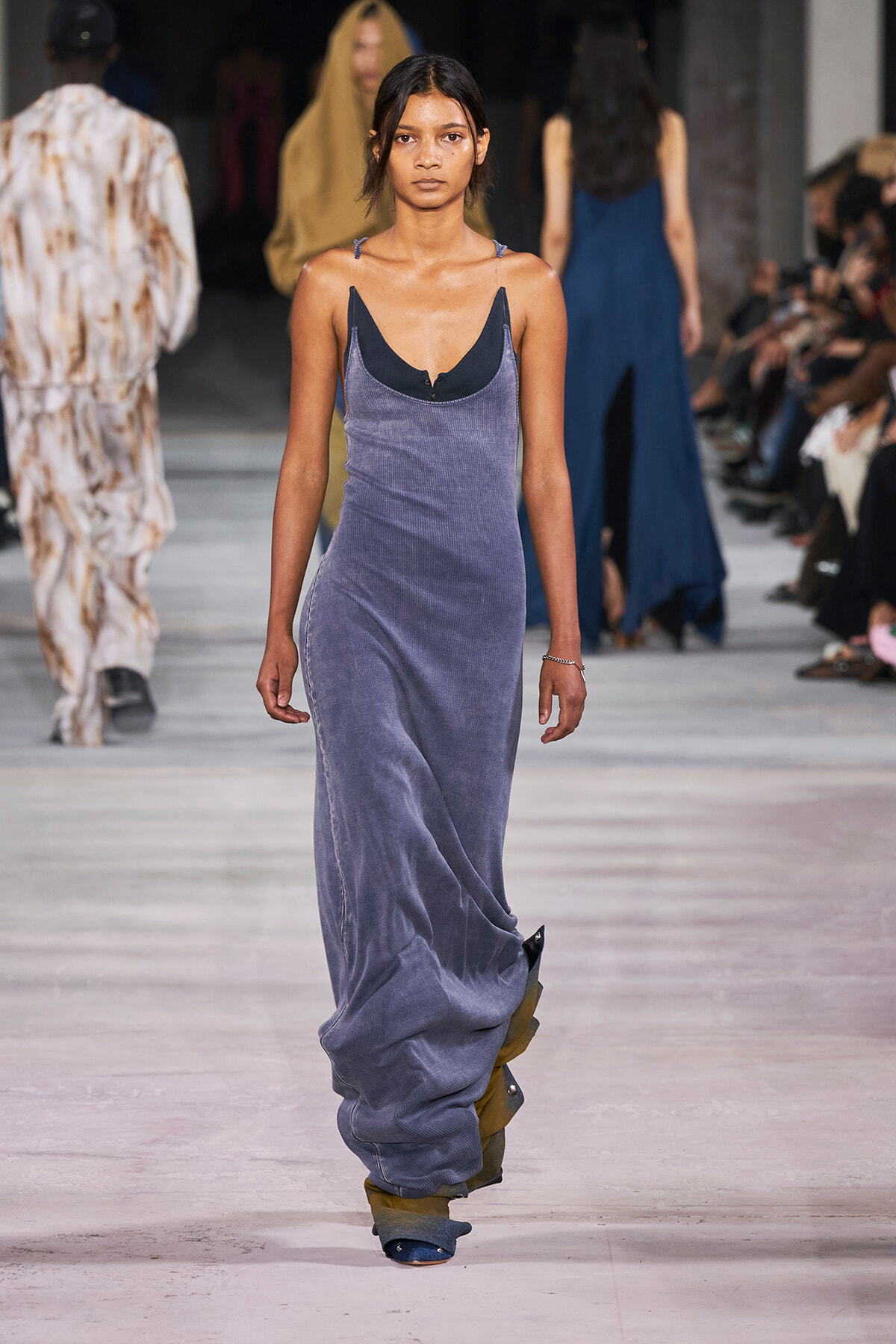 Female model walks the runway in a long lavender velvet dress with a deep neckline, audience visible on sides.