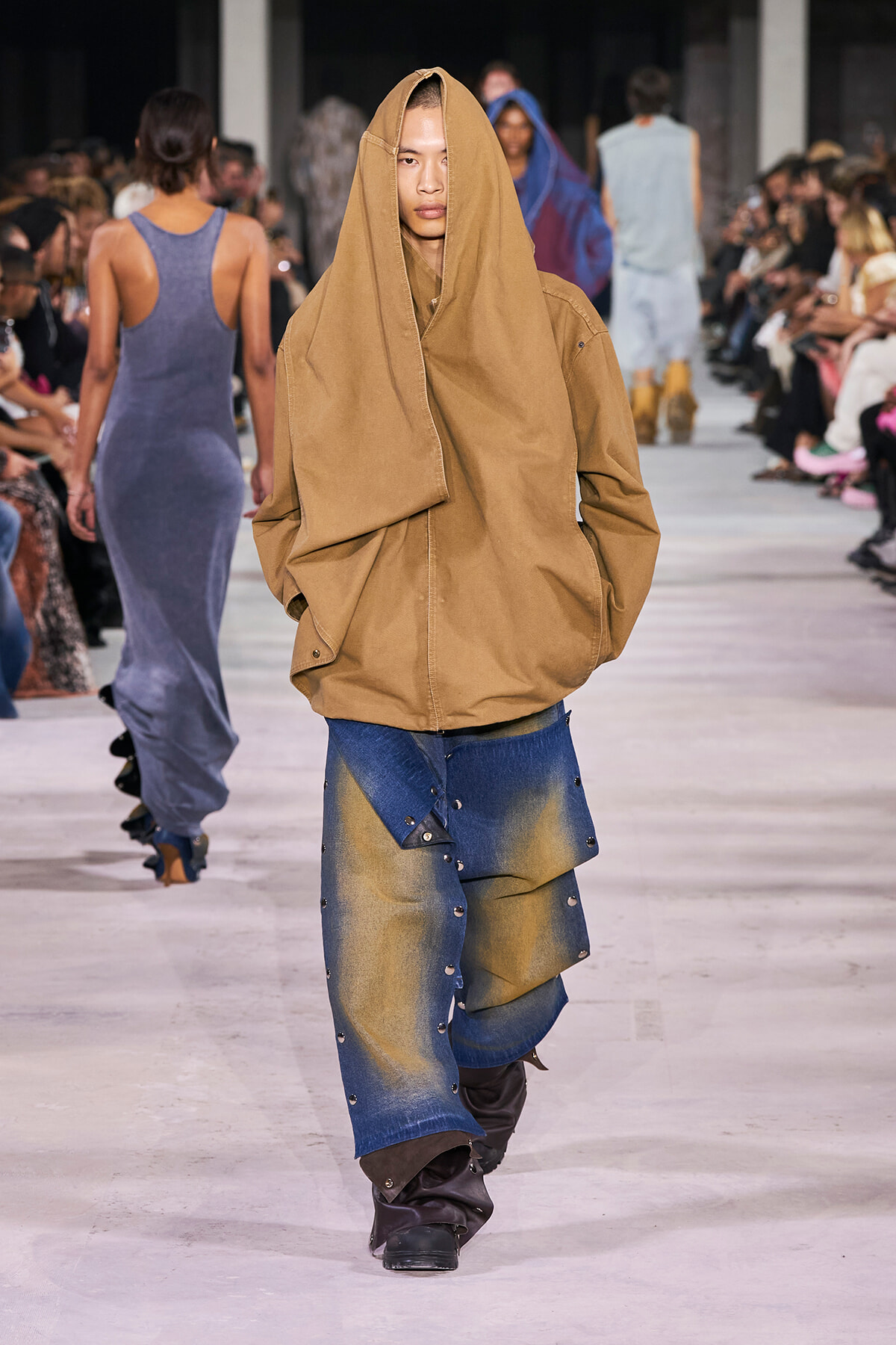 Runway model in an oversized tan hooded coat and patchwork denim pants walks the catwalk as seated audience watch