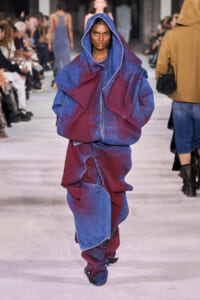 Model walking a fashion runway in a oversized blue and burgundy denim hooded outfit with hands in pockets; audience blurred in background.