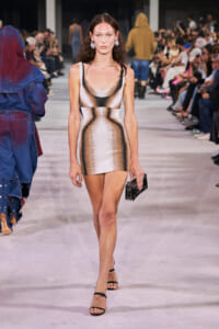 Model walks runway in a metallic gold-silver mini dress, black strappy heels, and a small black clutch; audience lines the catwalk.