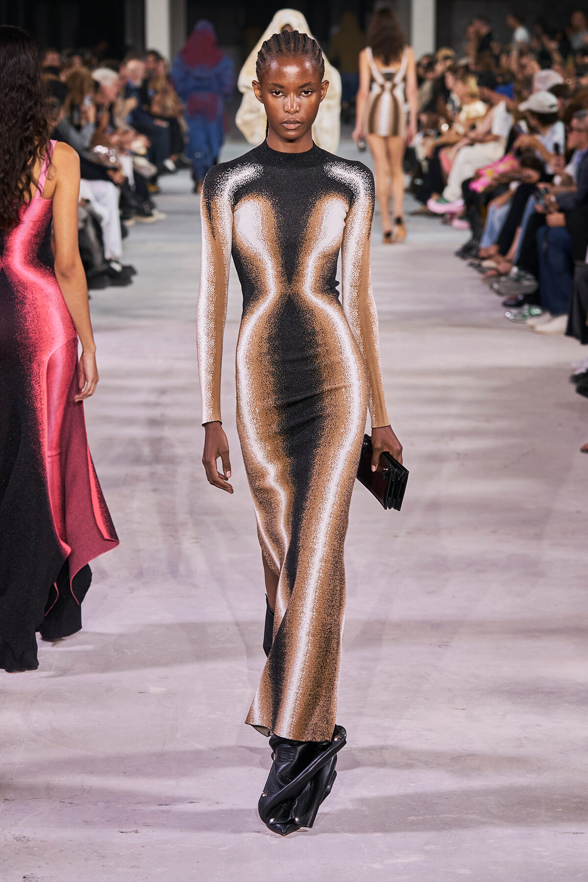 Model walking a fashion runway in a metallic gold and black form-fitting gown with a small black clutch.