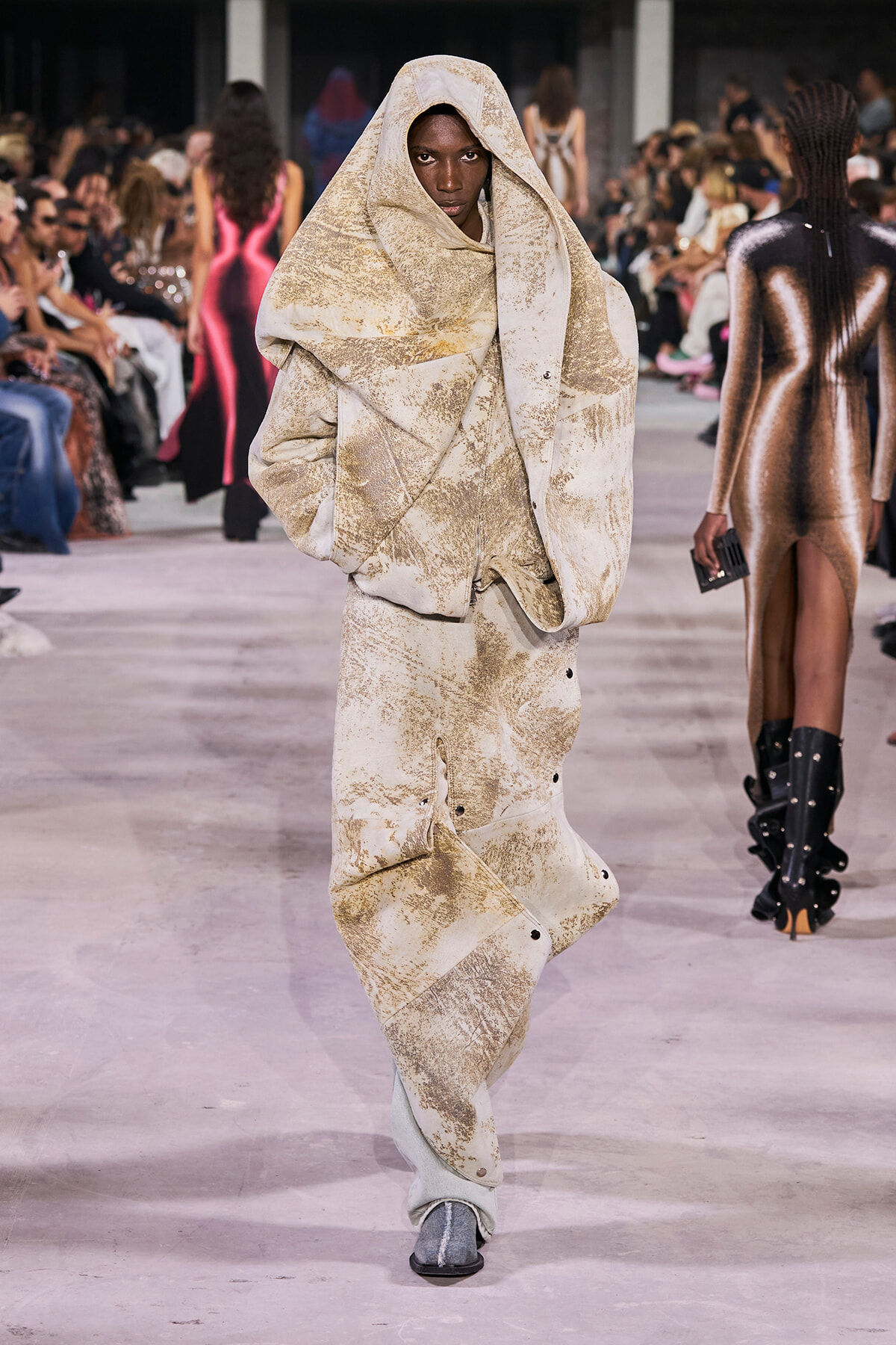 Model walks a fashion runway in a oversized beige textured coat with hood, audience on both sides watching