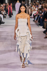 Model walking a fashion runway in a strapless tiered denim dress with large ruffles and white sandals, audience on either side.