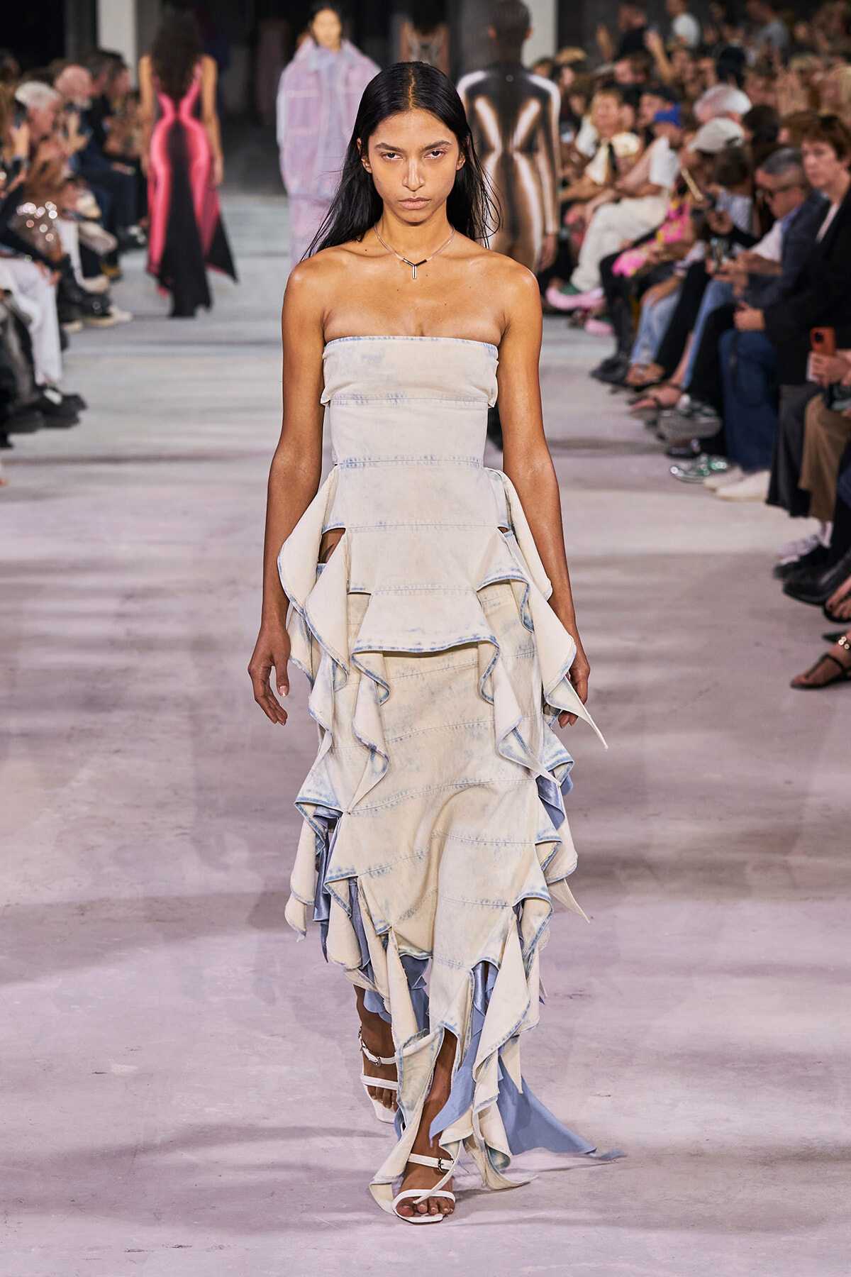 Model walking a fashion runway in a strapless tiered denim dress with large ruffles and white sandals, audience on either side.