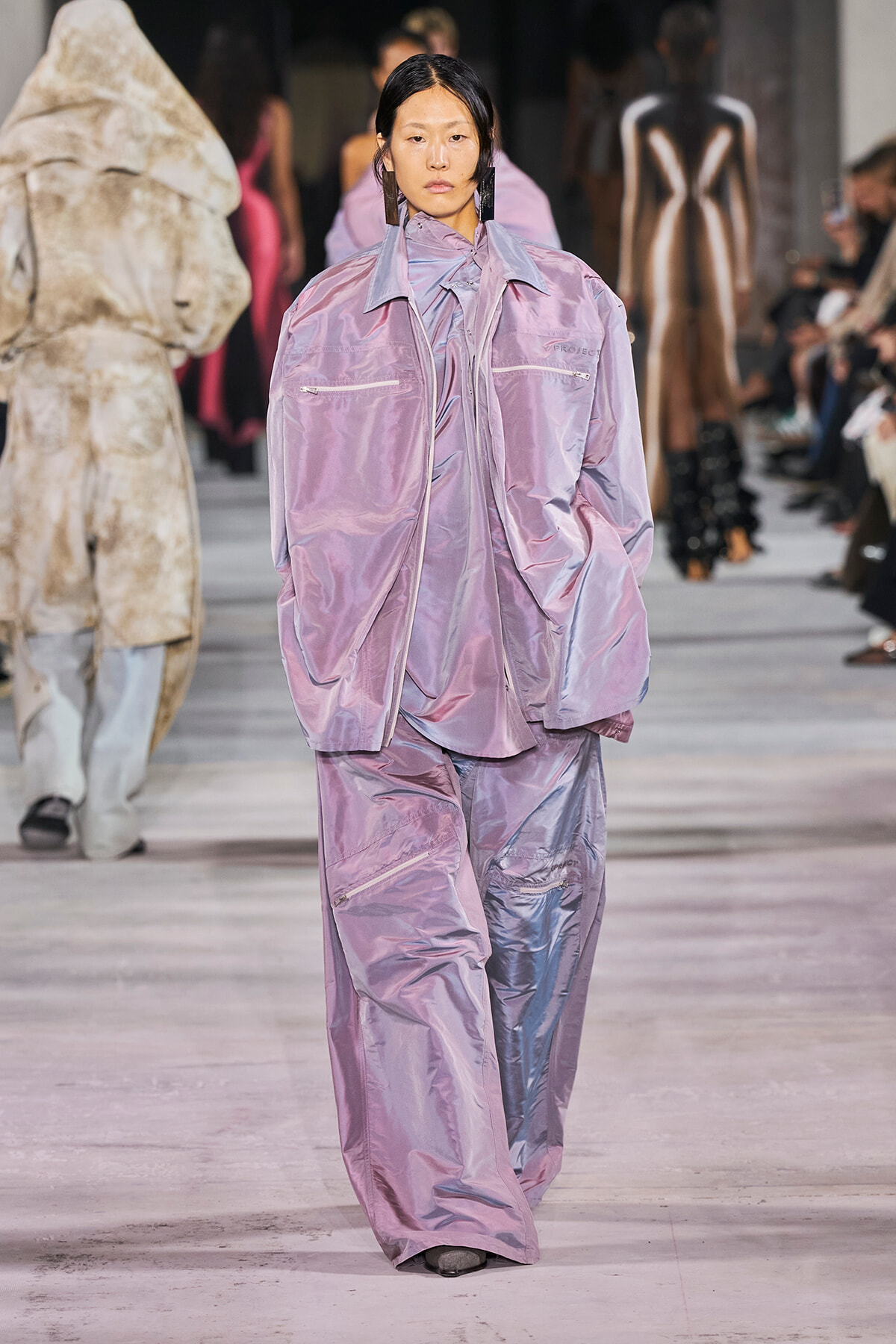 Model on a fashion runway wearing a loose, shiny lavender pantsuit with an oversized jacket in a soft lilac tone.