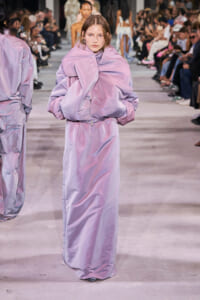 Model walks the runway in a lavender oversized puffer coat with a matching long dress; audience lined the catwalk in the background.