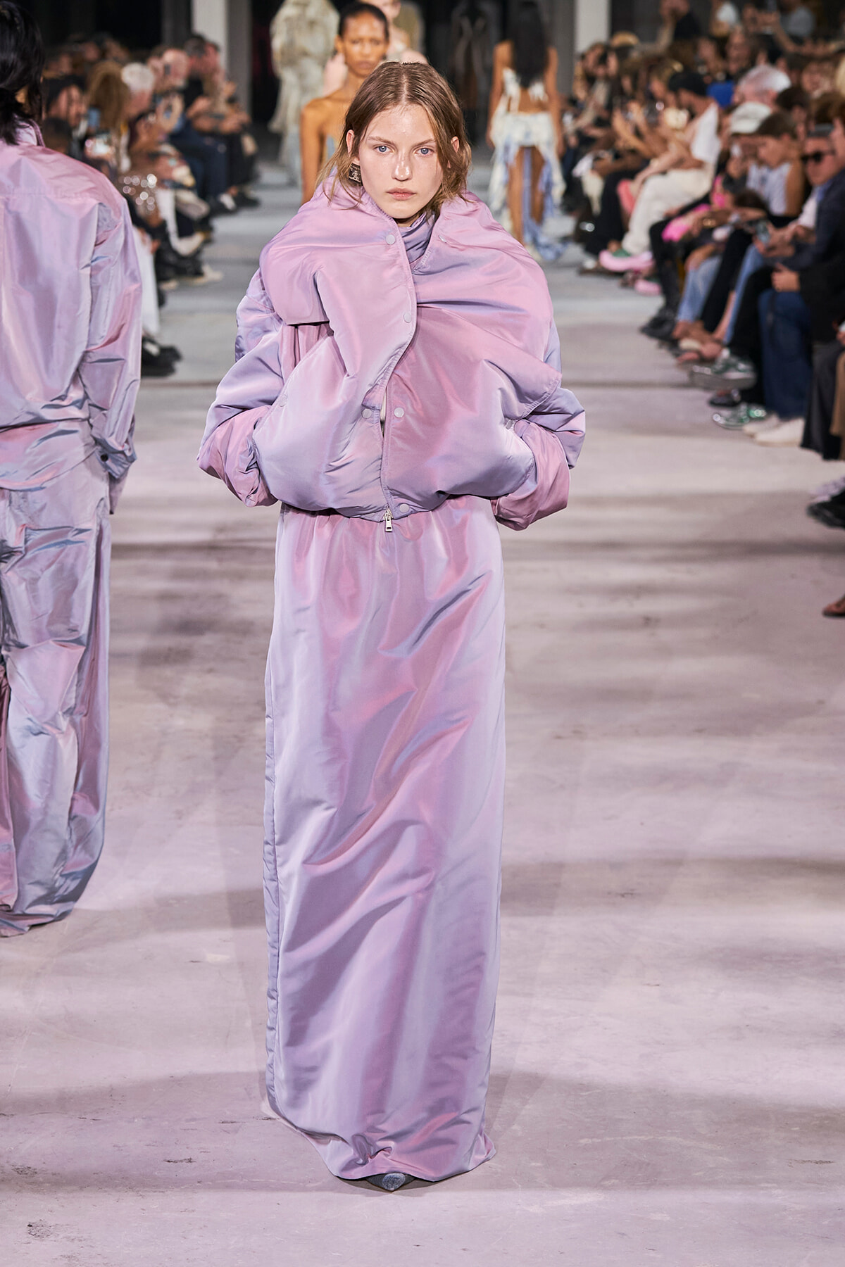 Model walks the runway in a lavender oversized puffer coat with a matching long dress; audience lined the catwalk in the background.