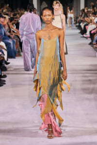 Model walks the runway in a metallic tiered dress in pastel gold, blue, and pink; audience watches from both sides.
