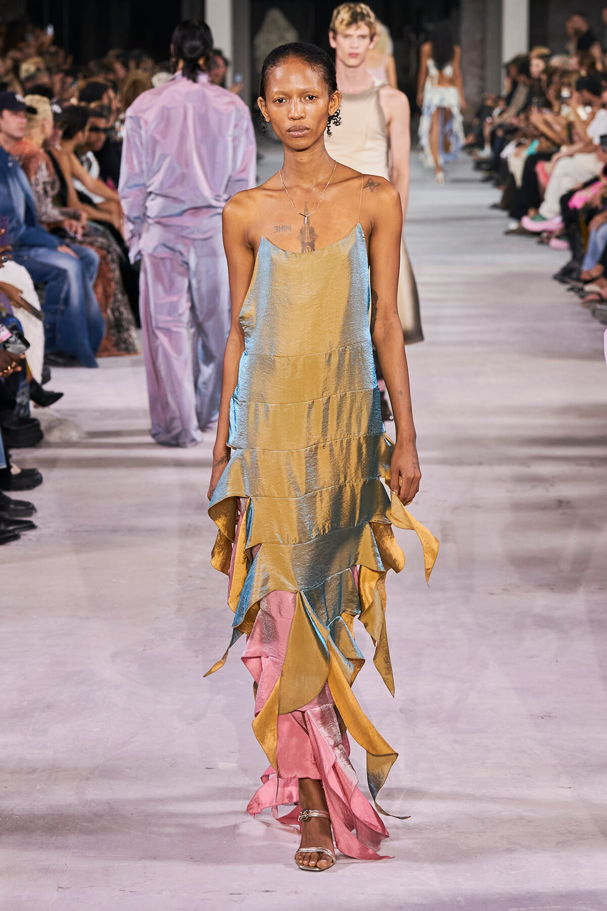 Model walks the runway in a metallic tiered dress in pastel gold, blue, and pink; audience watches from both sides.