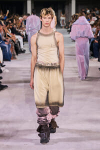 Male model walks a fashion runway in a beige satin tank top and gradient shorts, with spectators on both sides. Edit: belt and pink-toned wraps visible around the legs.