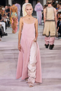 Bald model wearing a long pink dress with a side pocket on a fashion runway, audience in background