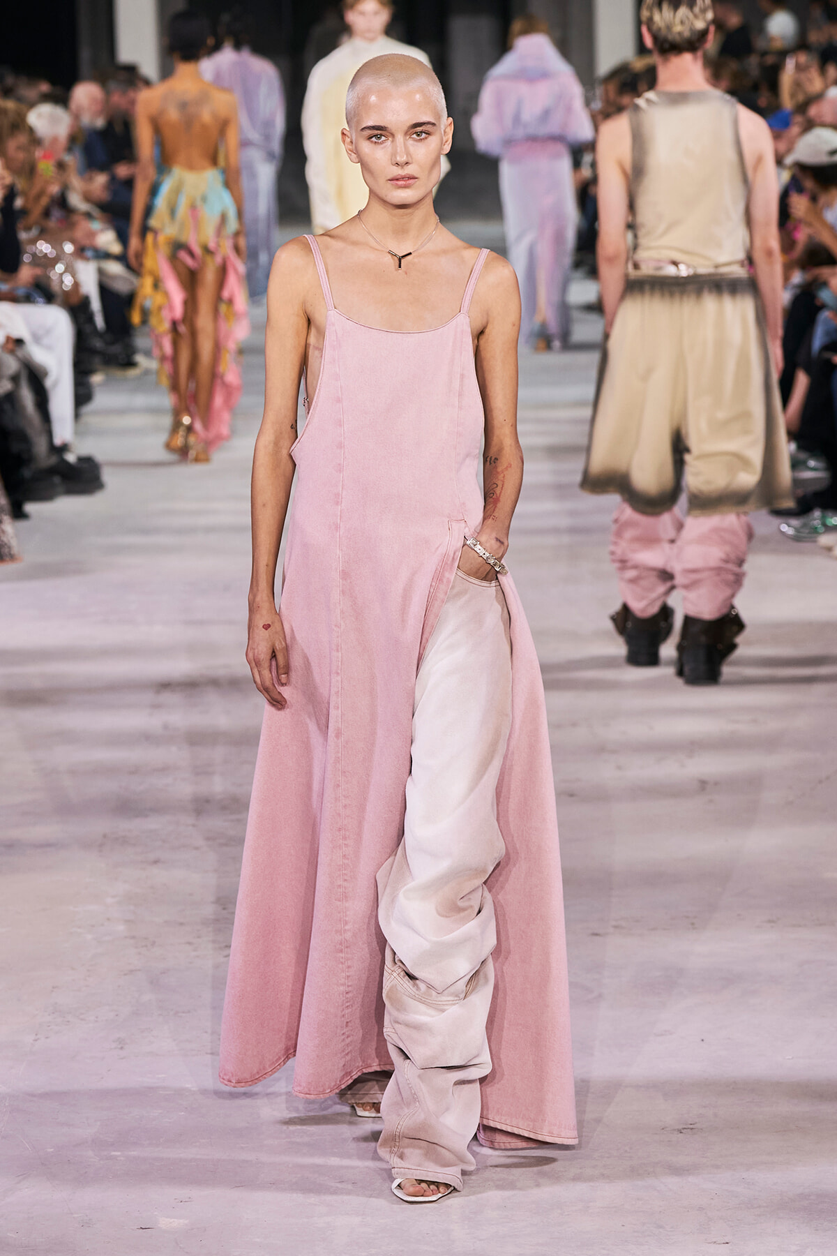 Bald model wearing a long pink dress with a side pocket on a fashion runway, audience in background