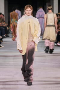 Model walks the runway in a pale yellow oversized coat over a sheer beige top and pink gradient pants? No, replace with: Model walks the runway wearing a pale yellow oversized coat over a light top and pink gradient pants.