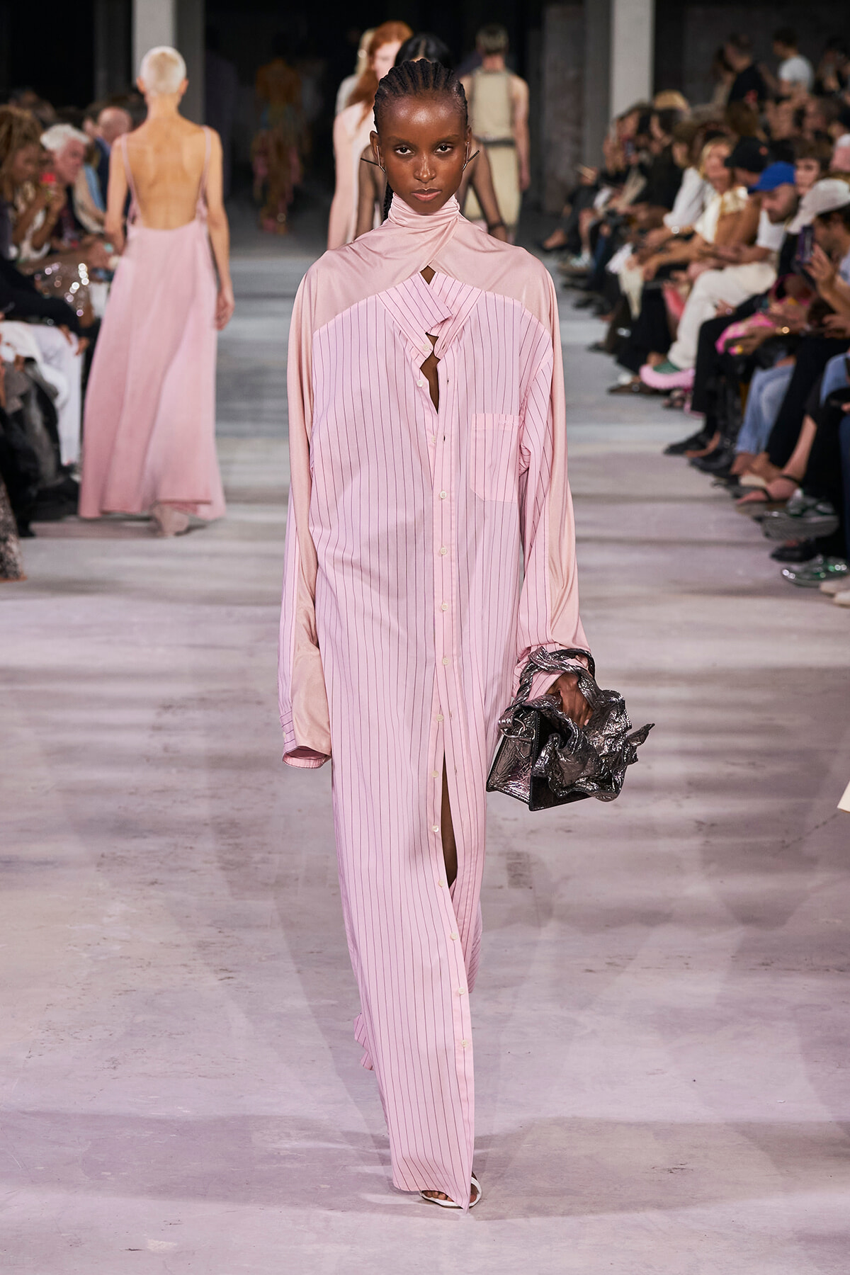 Model walks the runway in a pale pink, vertically striped shirt dress with a high-neck overlay, holding a crinkled metallic handbag.