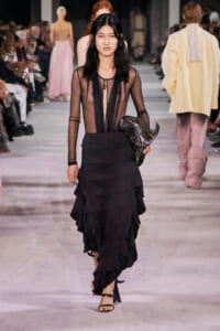 Female model walking a runway in a sheer black top with a tiered black skirt, holding a metallic jacket.