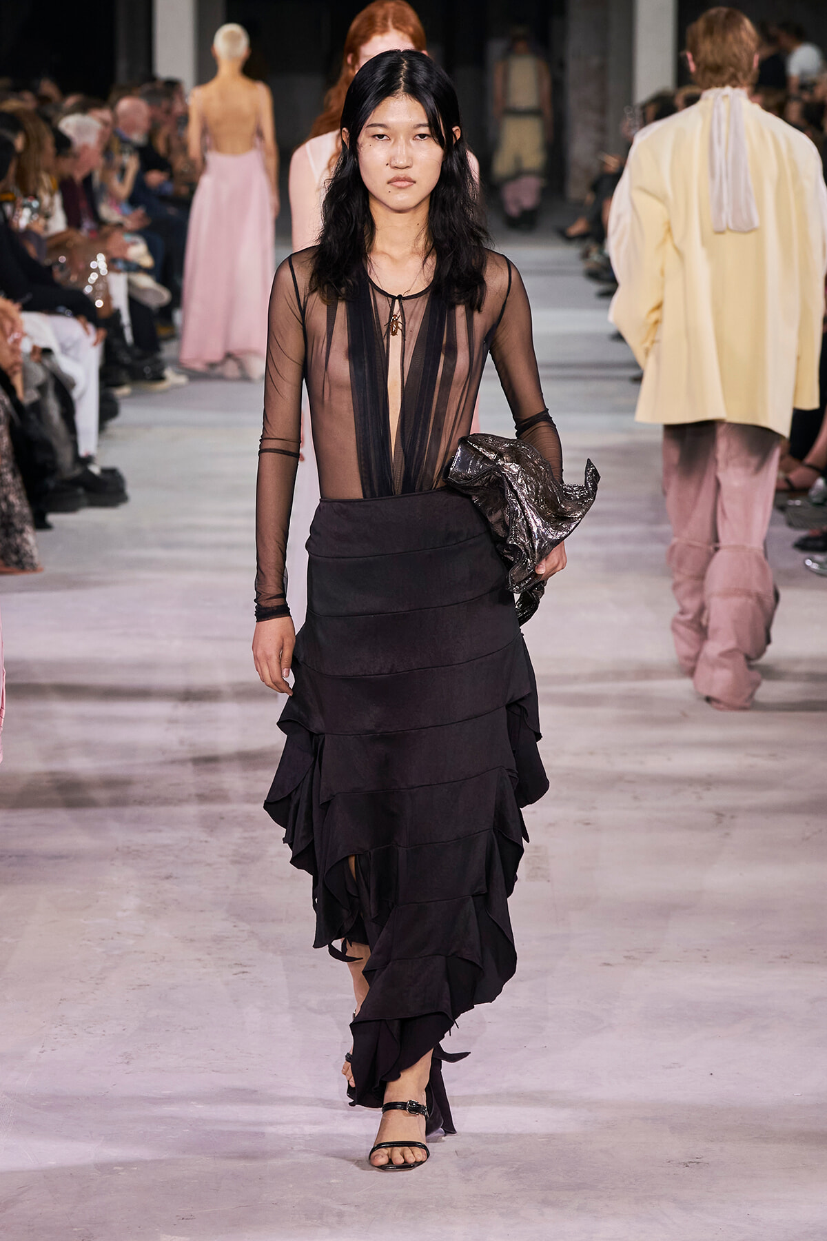 Female model walking a runway in a sheer black top with a tiered black skirt, holding a metallic jacket.