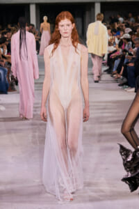 Model walks the runway wearing a sheer, pale pink-white gown that reveals undergarments, as spectators watch from the sides.