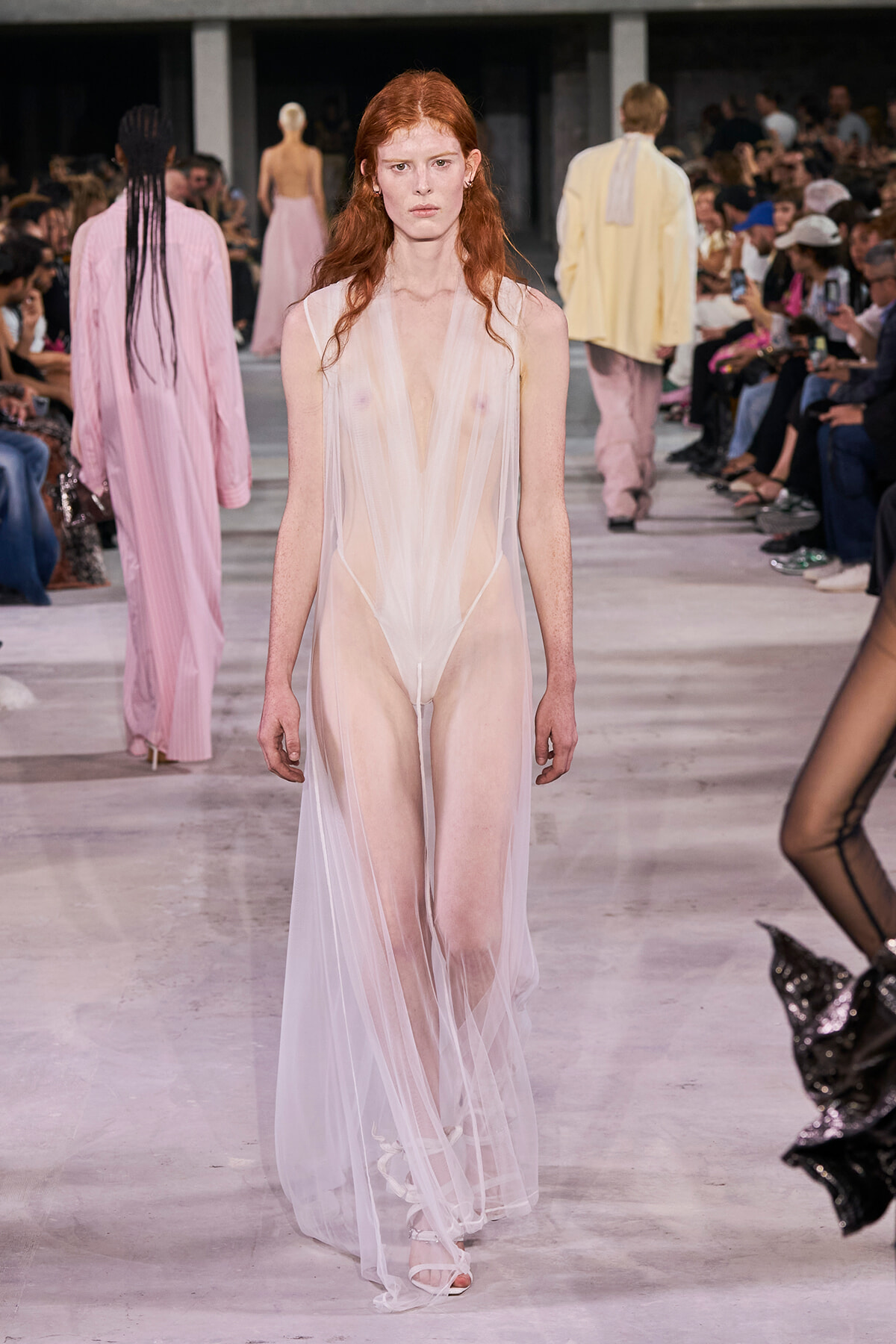 Model walks the runway wearing a sheer, pale pink-white gown that reveals undergarments, as spectators watch from the sides.