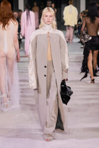 Model walking a fashion runway in a neutral-toned, oversized beige coat over a pale top and matching pants, carrying a black bag.
