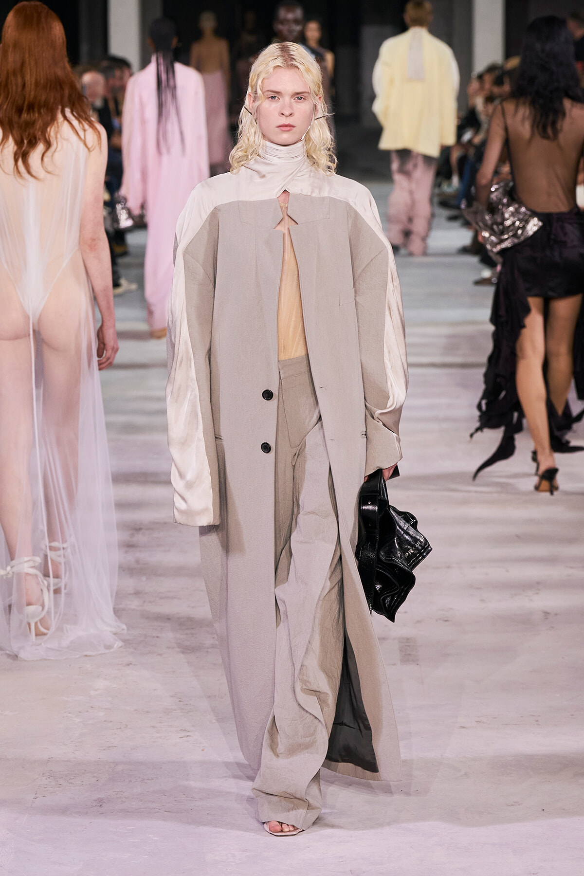 Model walking a fashion runway in a neutral-toned, oversized beige coat over a pale top and matching pants, carrying a black bag.
