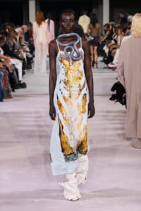Model walks the runway wearing a pale blue gown with a gold-yellow abstract pattern and a gray snake accessory draped around her neck.