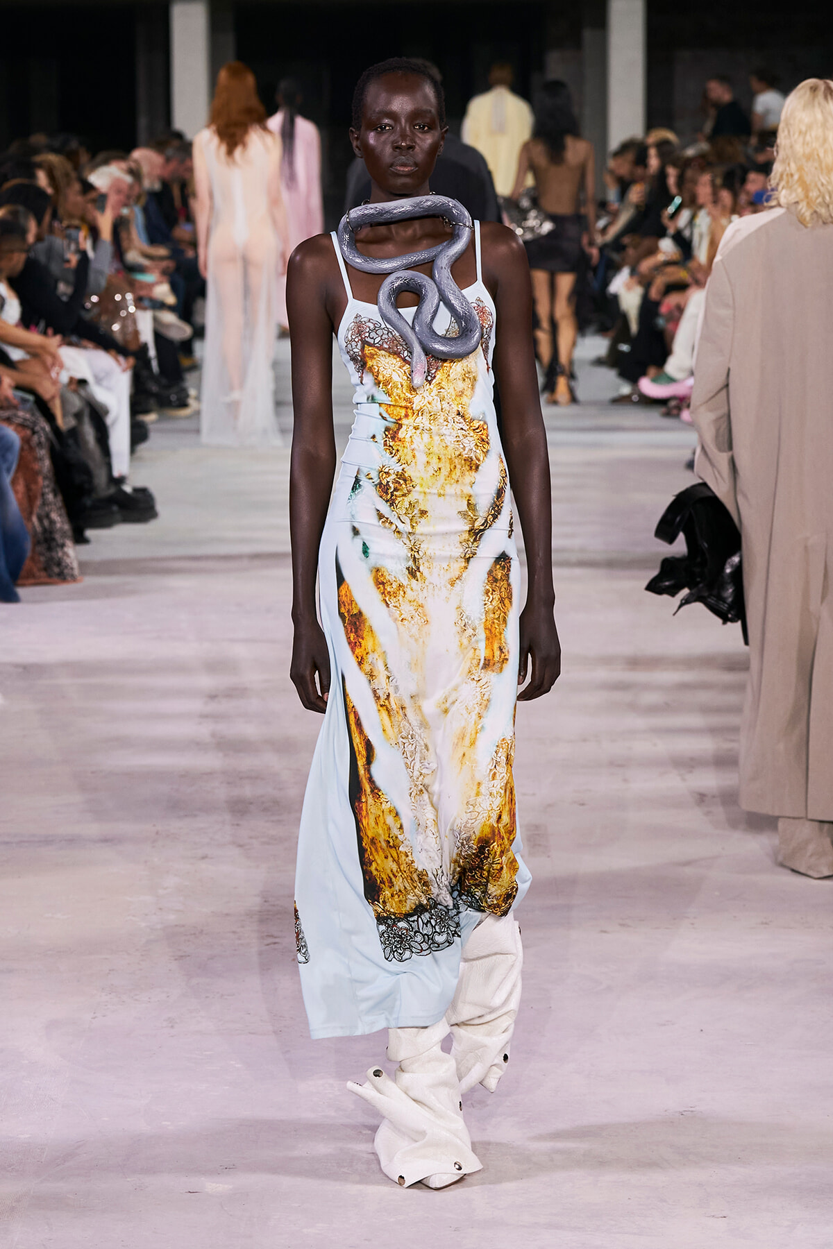 Model walks the runway wearing a pale blue gown with a gold-yellow abstract pattern and a gray snake accessory draped around her neck.
