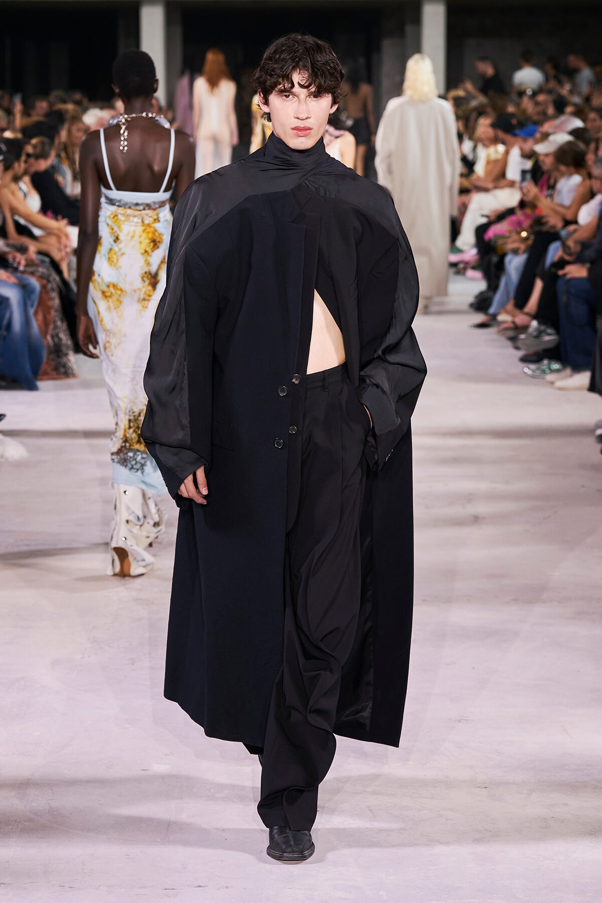 Male model in a long, oversized black coat with a high neck, walking a fashion runway; dramatic, minimal styling with a partially open front revealing a pale chest. Audience seated along the runway in the background.
