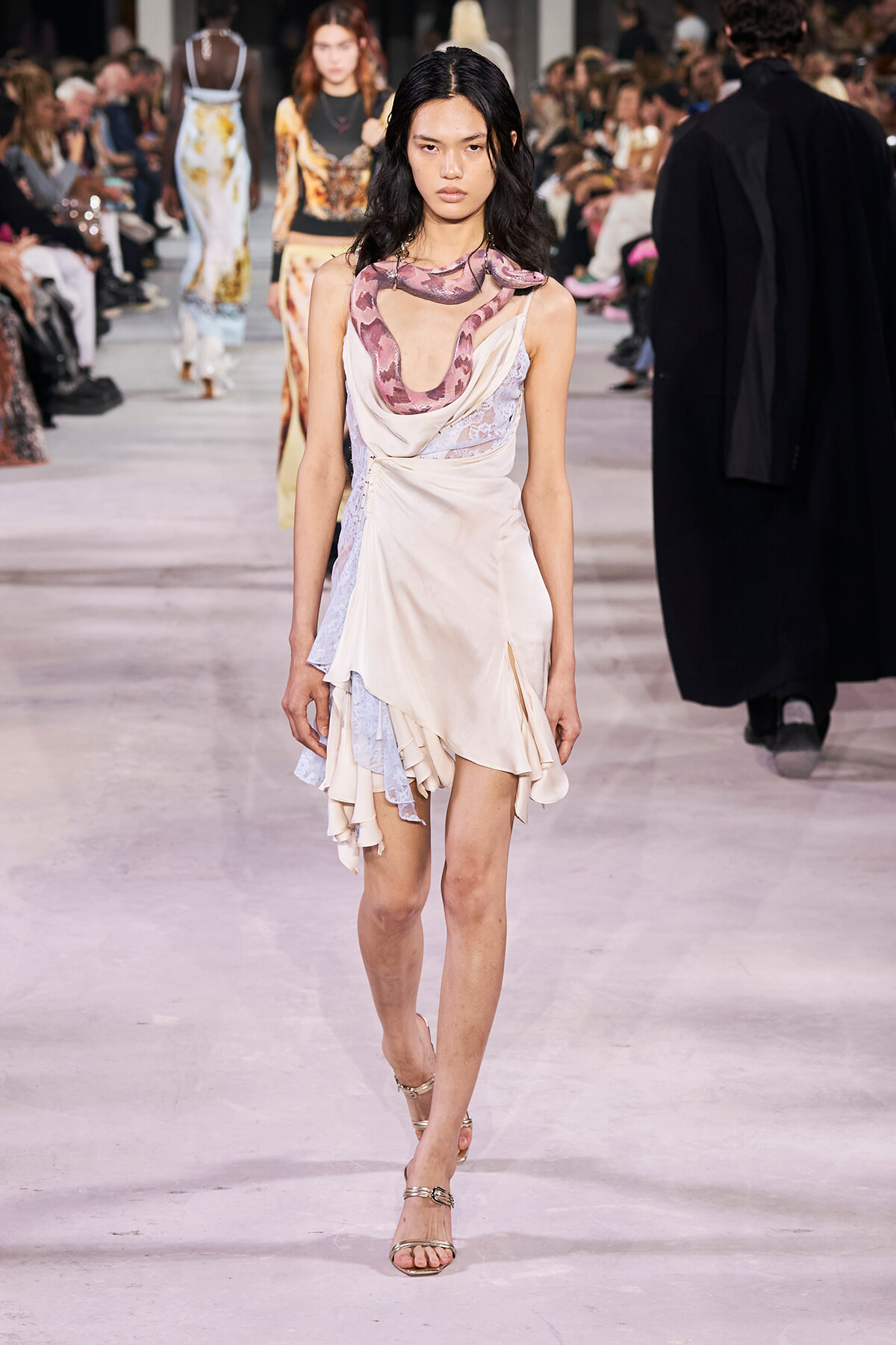 Model walks a fashion runway in a pale cream draped satin dress with lilac lace panels and a pink snake accessory around the shoulders; audience in background.