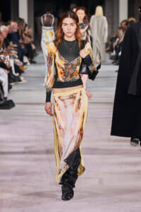 Model walking a fashion show runway in a gold, black print long-sleeve top with matching printed pants, carrying a black bag on her shoulder