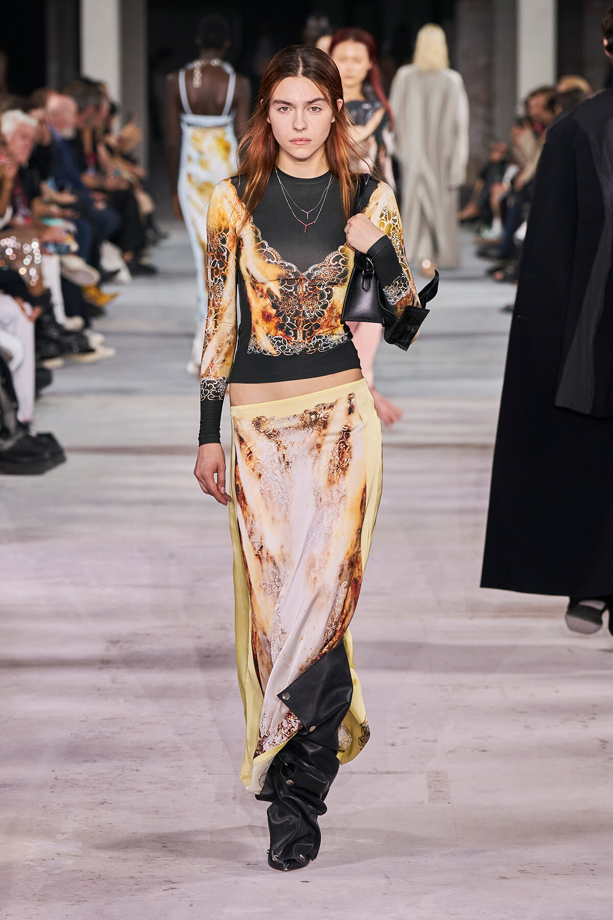 Model walking a fashion show runway in a gold, black print long-sleeve top with matching printed pants, carrying a black bag on her shoulder