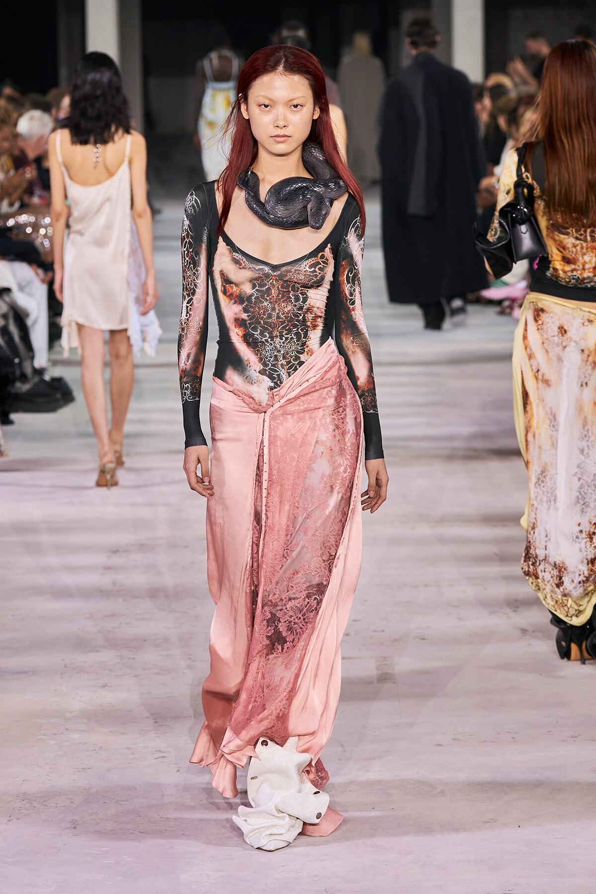 Model walking a runway in a pink satin lace dress with a black snake accessory around her neck, audience in background.