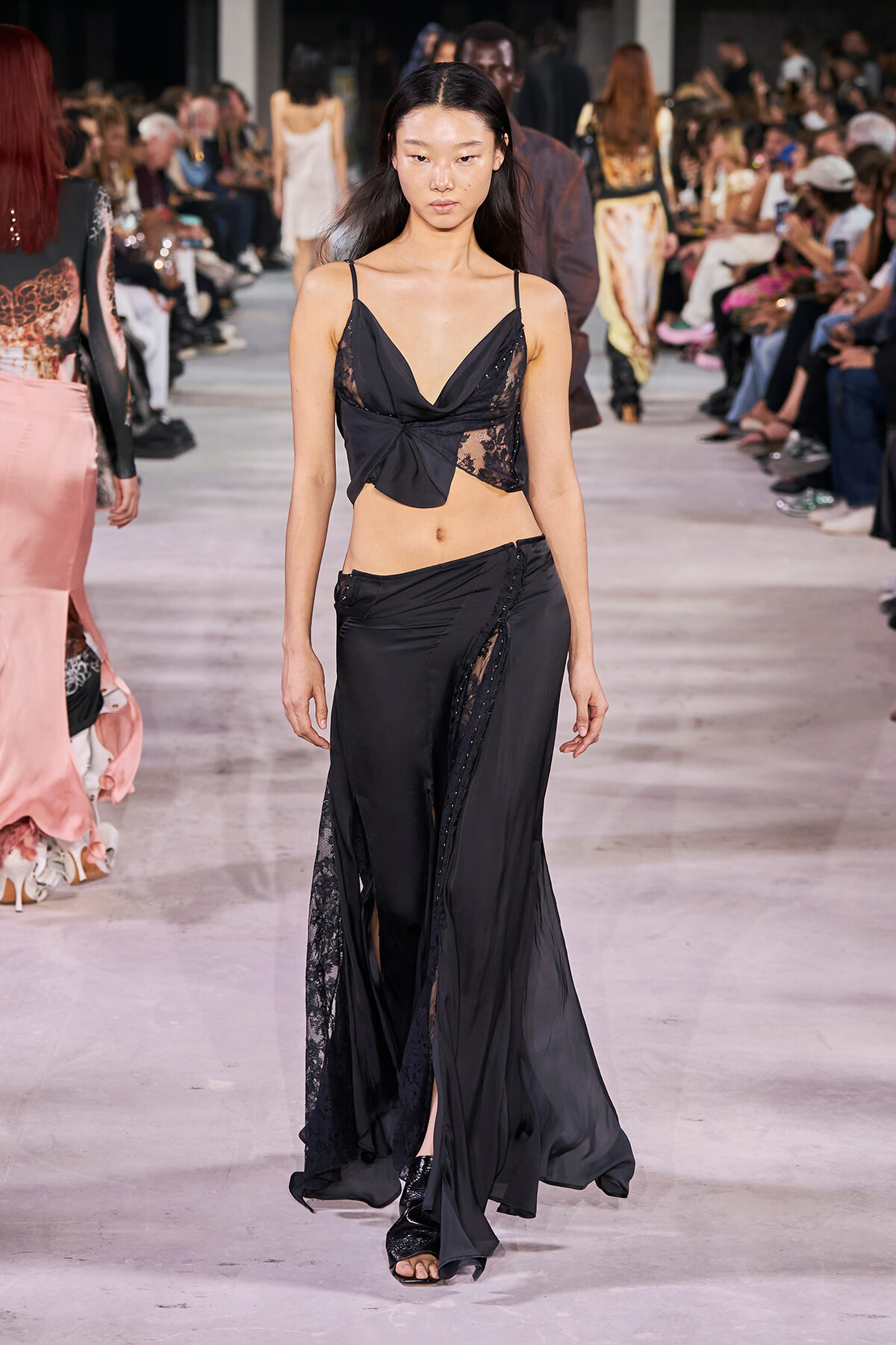 Model walks the runway wearing a black, asymmetrical, lace-paneled crop top and matching floor-length skirt with slits, during a fashion show.
