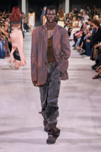 Male runway model with dark skin wearing a rust-colored oversized blazer over a brown shirt and distressed jeans walks the catwalk as a blurred audience watches from both sides.