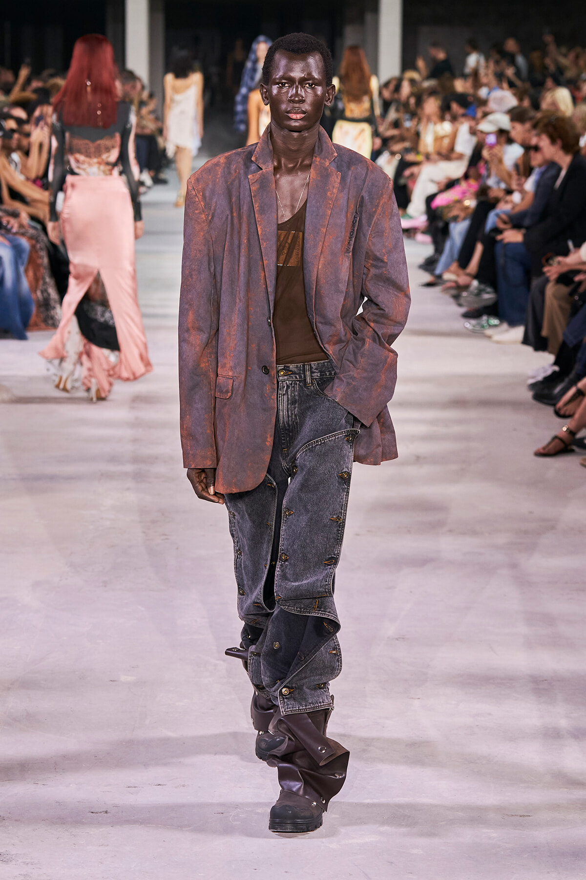 Male runway model with dark skin wearing a rust-colored oversized blazer over a brown shirt and distressed jeans walks the catwalk as a blurred audience watches from both sides.