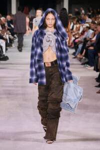 Runway model walking in a blue plaid oversized hooded jacket over a gray knotted top and brown cargo pants, audience in the background