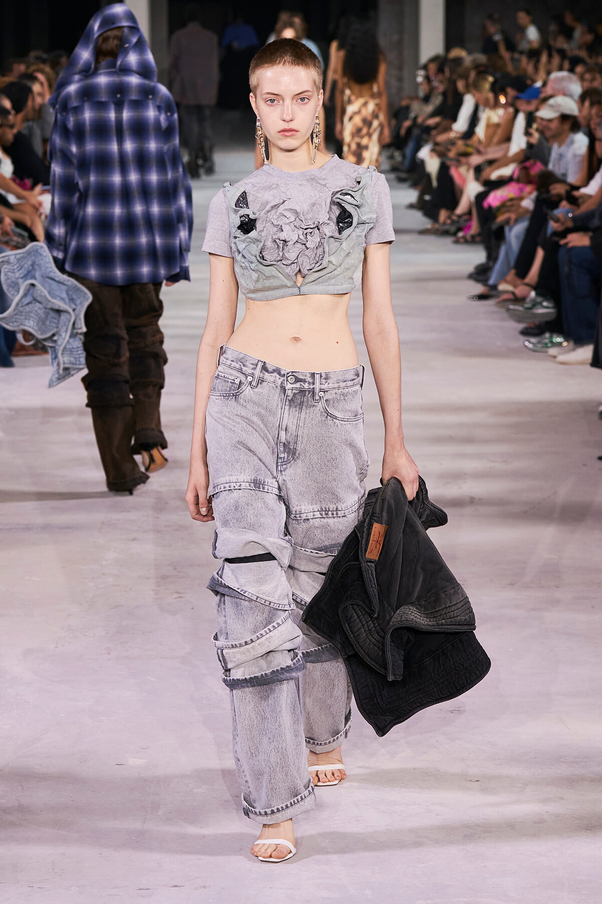Model walking a runway in a cropped gray top with fabric rosettes and oversized washed jeans, carrying a black jacket.
