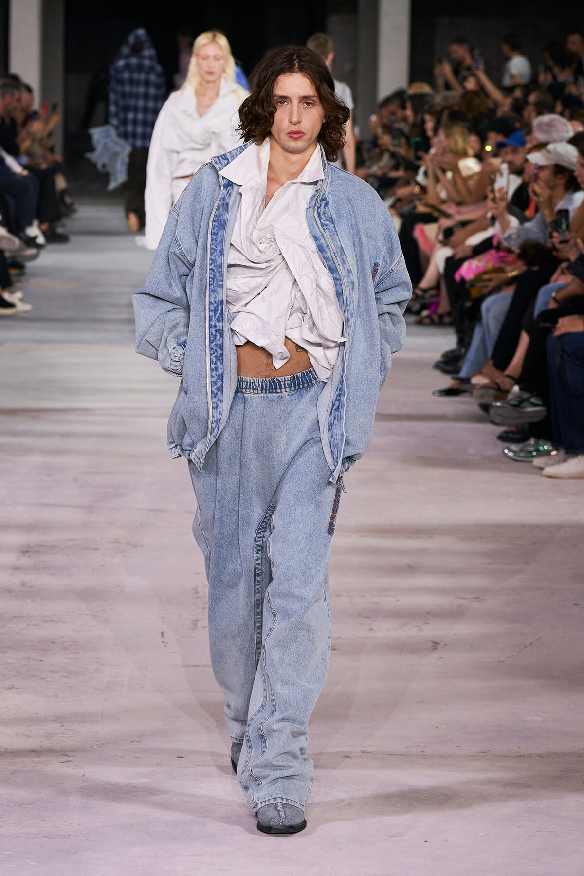 Male model walks a fashion runway in an oversized light denim jacket and matching jeans, with a crumpled white top, as the audience looks on.