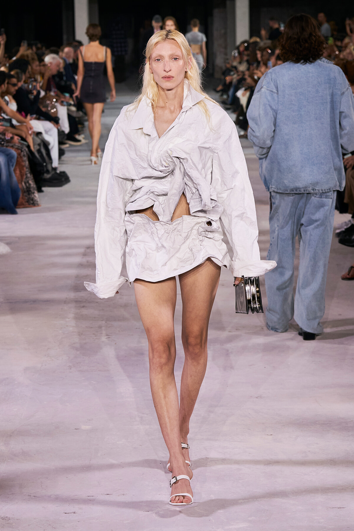 Female model walks a runway in a crumpled white oversized top tied at the waist, with a matching bottom, and a small metallic clutch in hand.