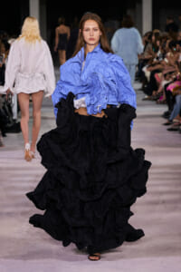 Model walks the runway in a blue striped oversized shirt and a dramatic black ruffled skirt.
