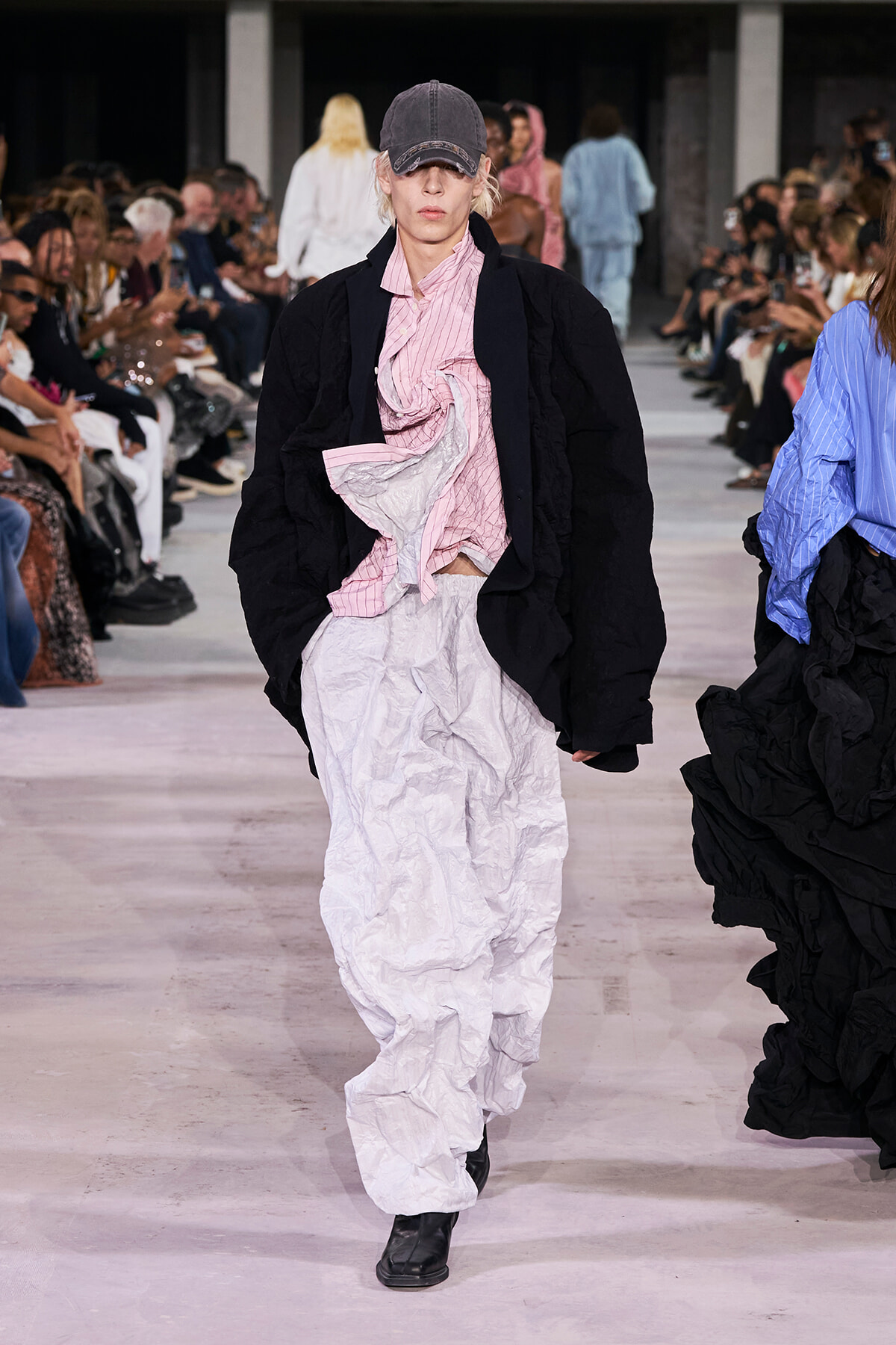 Model walks the runway in a black oversized jacket over a pink crinkled blouse with a voluminous white crinkled skirt; gray baseball cap and black boots; audience on both sides observing.