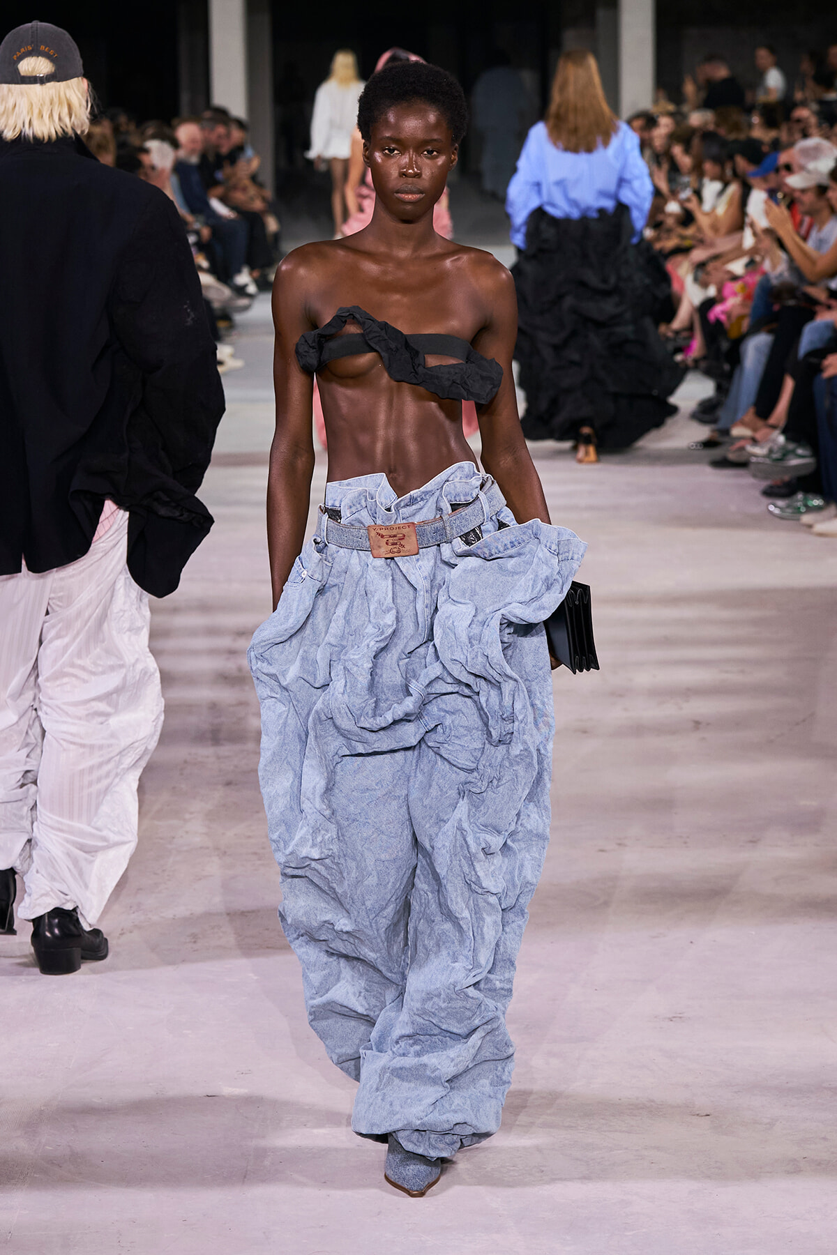 Model walks the runway in an avant-garde denim ensemble with a crumpled light-blue pant look and a black twisted strap across the chest, audience in background.